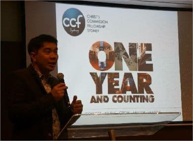 A speaker addressing a CCF Sydney gathering in front of a banner reading One Year and Counting